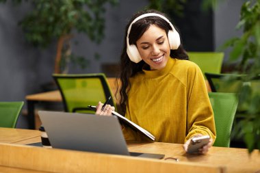 Neşeli kadın çalışma alanında dizüstü bilgisayar ve akıllı telefonla masasında birden fazla iş yapıyor..