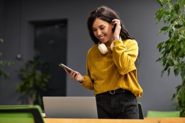 Modern bir ofiste, genç ve zarif bir kadın telefonunu kullanarak bir dizüstü bilgisayar üzerinde çalışırken gülümser..