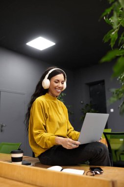 Gündelik kıyafetli zarif bir kadın modern bir ofiste dizüstü bilgisayarla çalışırken gülümsüyor..