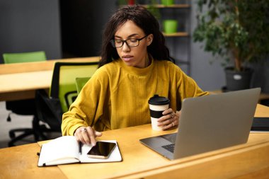 Güzel genç bir kadın şık bir ofis ortamında kahvesini yudumlarken işine odaklanıyor..