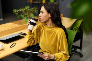 Genç profesyonel, hayat dolu bir ofis ortamında kahvesini yudumlarken düşünceli bir şekilde yansıtır..