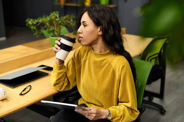 Young professional reflects thoughtfully while sipping coffee in a lively office environment.
