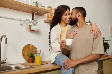 Bir çift mutfakta birlikte oturur, gülümserler ve içkilerinin tadını çıkarırken şefkatli bir öpücük paylaşırlar..