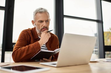 Ciddi bir adam, çağdaş bir ofis ortamında dizüstü bilgisayarı üzerinde çalışırken not alır..