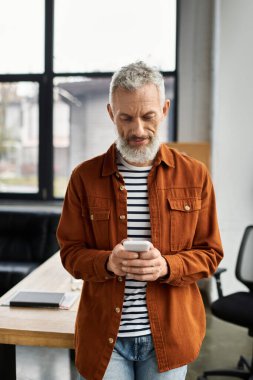 Çağdaş bir ofis masasında akıllı telefon kullanan gri saçlı ve sakallı biri..