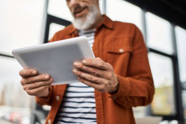 Sakallı yaşlı bir adam, çağdaş bir ofiste dururken tabletle etkileşime geçiyor..