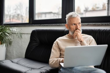 Düşünceli bir birey, modern bir ofis ortamında, bir dizüstü bilgisayarda çalışmaya dalmış bir şekilde bir koltuğa oturur..