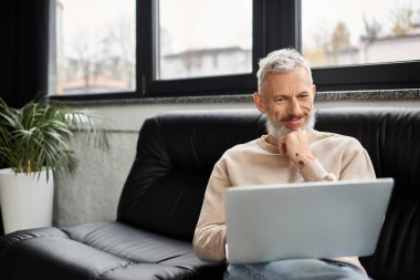 Sakallı adam, çağdaş bir ofiste koltukta oturur ve laptopuyla düşünceli bir şekilde meşgul olur..