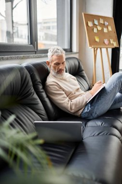 Gri saçlı bir adam, parlak, rahat bir ofiste dizüstü bilgisayara notlar yazar..