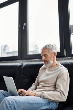 Yaşlı bir adam modern bir ofiste dizüstü bilgisayarını büyük pencerelerden kullanır..