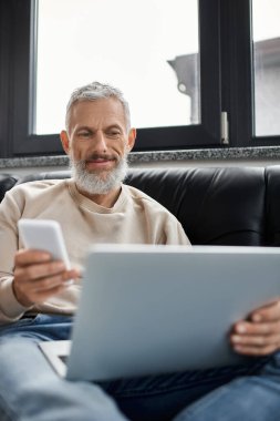 Sakallı bir adam dizüstü bilgisayarında yazı yazıyor. Akıllı telefon elinde. Ofis ortamının tadını çıkarıyor..