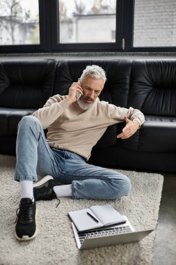 Gri saçlı bir adam ofisinin zemininde oturup telefonla konuşuyor..