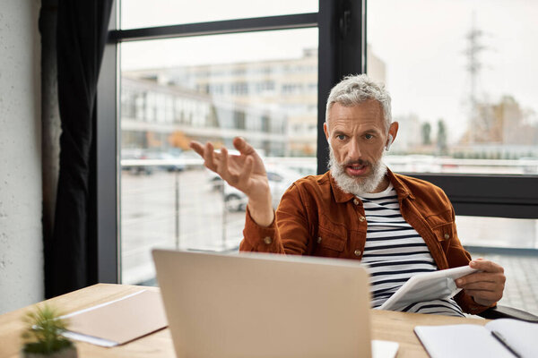 A man passionately expresses his thoughts while seated at a modern desk, surrounded by paperwork.