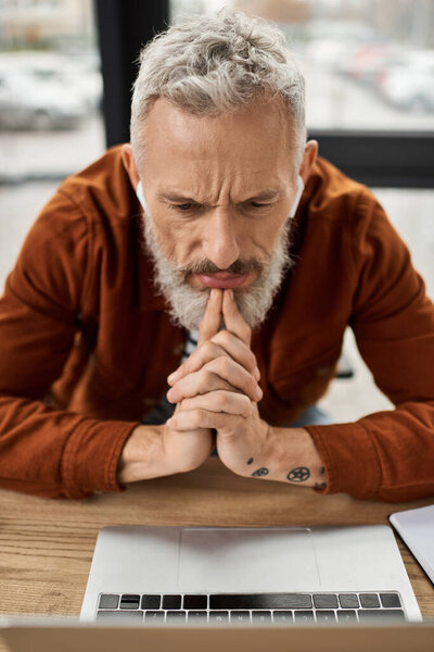 Вдумчивый человек размышляет над ноутбуком в гладком офисе с естественным светом.