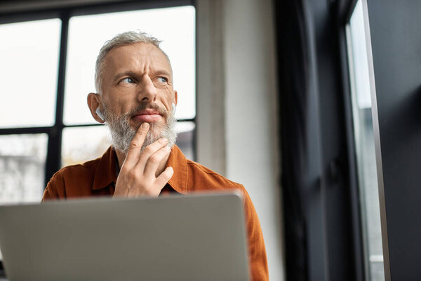 A man thoughtfully considers his work while seated at a laptop in a bright office.