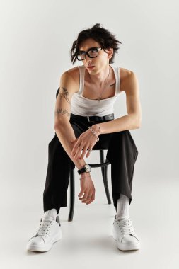 Beyaz kolsuz tişörtlü genç adam kendinden emin poz veriyor, modaya uygun görünüşünü ve kendini ifade edişini vurguluyor..