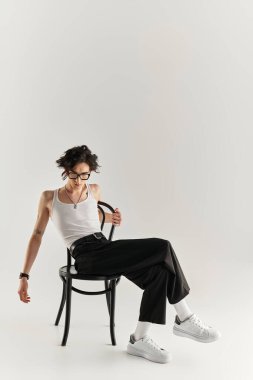 Beyaz kolsuz tişörtlü zayıf bir genç otururken moda tarzını sergiliyor.