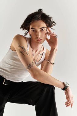 Beyaz kolsuz tişörtlü genç bir adam şık bir poz veriyor, stüdyoya güven saçıyor..