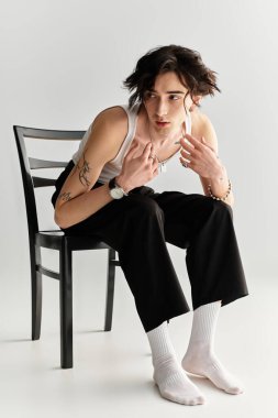 Sandalyeye oturan sıska genç adam kendine güvenen ve tarz sahibi bir görünüm sergiliyor.