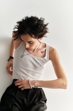 Beyaz kolsuz tişörtlü zayıf bir genç, sade bir arkaplana karşı güvenle poz verir..