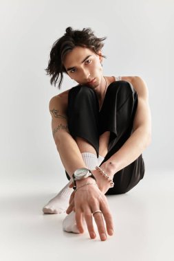 Şık bir genç adam stüdyo ortamında rahat bir şekilde oturuyor ve modaya uygun kıyafetlerini sergiliyor..