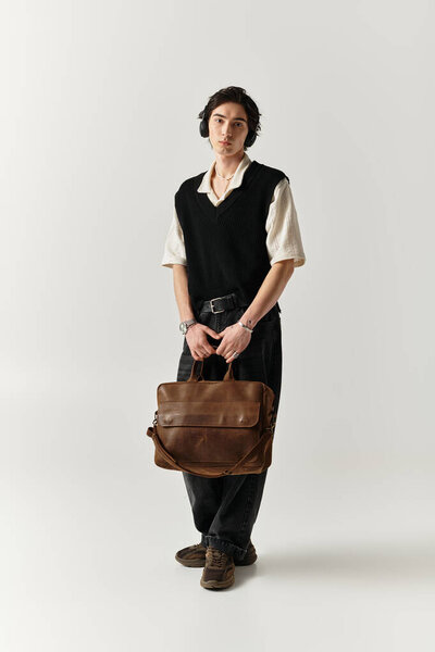 Красивый молодой человек уверенно стоит в студии в стильном наряде и с сумкой в руках..