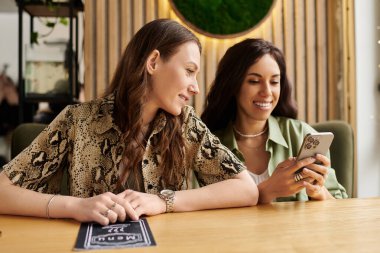 İki kadın bir kafede aynı telefonu paylaşırken birlikte gülüp gülümsüyor..