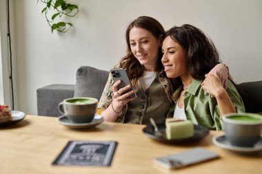 Neşeli bir lezbiyen çift, kafede akıllı telefona bakarken yeşil çay eşliğinde kahkahalarını paylaşıyor..