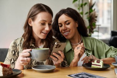 İki kadın neşeli bir anı paylaşıyor, kahve ve tatlıdan keyif alırken akıllı telefon kullanıyorlar..