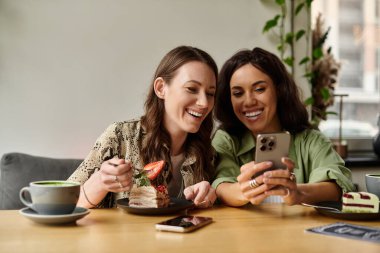 Neşeli bir lezbiyen çift, bir kafede tatlıdan zevk alıyor, akıllı bir telefonla eğlenirken gülüyorlar..