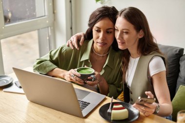 İki kadın yan yana oturur, içkilerini paylaşır ve neşeli bir kafe ortamında gülümserler..