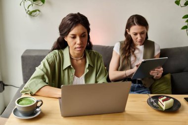 İki kadın bir kafede rahat bir anı paylaşıyor, biri dizüstü bilgisayara odaklanmış, diğeri de not alıyor..