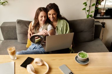 İki kadın, rahat bir kafede dizüstü bilgisayarla çalışırken yan yana oturup, gülümsüyor ve tatlının tadını çıkarıyor..