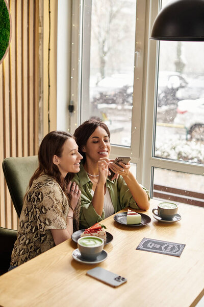 Two women smile together while enjoying coffee and delicious desserts in a warm cafe.