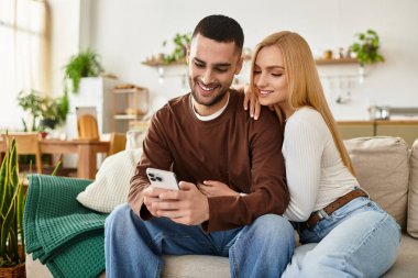 Bir çift, rahat bir oturma odasında akıllı telefon kullanırken kanepede gülüp ısınıyor..