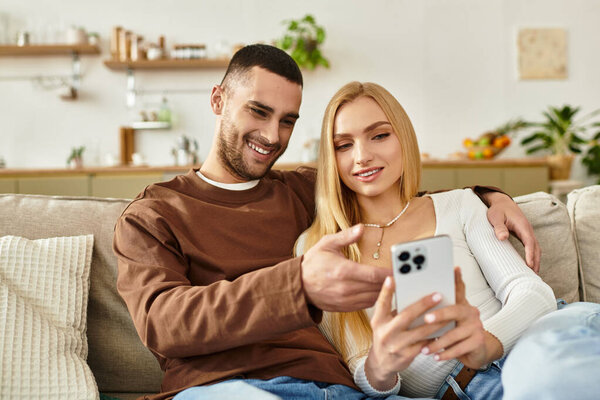 A couple sitting closely on a couch smiles joyfully together while sharing a phone.