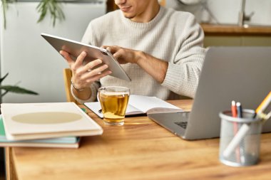 Tarz sahibi genç bir adam, sıcak bir ev ofisinin tabletine bakıyor, yanında çay ve not defteri var..