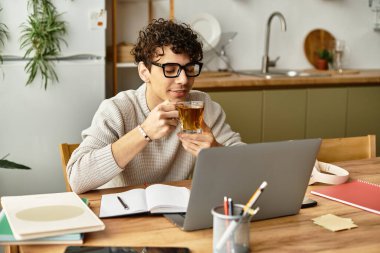 Yakışıklı genç adam çayını yudumluyor, rahat bir mutfak ortamında dizüstü bilgisayarına odaklanıyor.