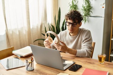 Süveter giyen genç bir adam masadaki dizüstü bilgisayarına odaklanırken kulaklığı ayarlıyor..