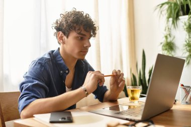 Genç adam rahat ve şık bir ortamda elinde kalem tutarken dizüstü bilgisayarına konsantre oluyor..