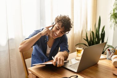 Yakışıklı genç adam telefonda konuşurken gülümsüyor ve güneşli bir masada dizüstü bilgisayarıyla çalışıyor..