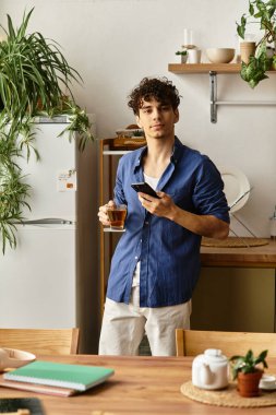 Yakışıklı genç adam bir fincan çay tutuyor ve canlı bir mutfak ortamında telefonuna bakıyor..