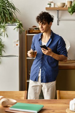 Yakışıklı genç adam iyi aydınlatılmış bir mutfakta elinde bir fincan çayla durup telefonuna bakıyor..