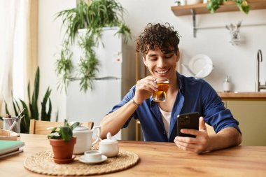 Genç, yakışıklı bir adam çayını yudumlarken gülümser ve rahat bir masada telefonunu kontrol eder..