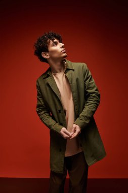 Kıvırcık saçlı genç adam şık bir palto giyiyor, kırmızıya karşı dururken düşünceli görünüyor..
