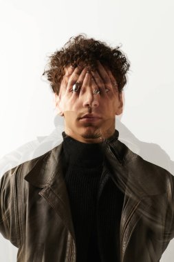 Genç ve yakışıklı bir adam, özgüven içinde kendine has tarzını sergileyerek sade bir arkaplanda duruyor..