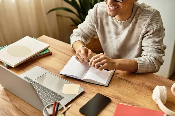 Genç bir adam gülümsüyor, renkli defterler ve teknoloji arasında düşünceli bir şekilde yazıyor..