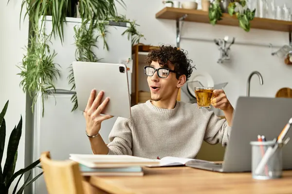 Kıvırcık saçlı genç bir adam, rahat bir çalışma alanında tablet kullanırken çaydan zevk alır..
