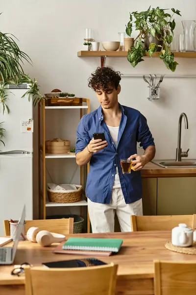 Gündelik giyinen yakışıklı genç adam modern bir mutfakta akıllı telefonunu kontrol ederken içki yudumluyor..