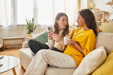 İki kadın kanepede rahatlar, sıcak bir içki içerler ve rahat yaşam alanlarında keyifli sohbetler ederler..